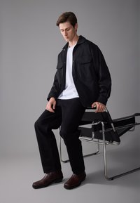 Veste noire légère avec deux poches avant, t-shirt blanc, pantalon noir et bottes en cuir marron. Assis sur une chaise moderne en métal.