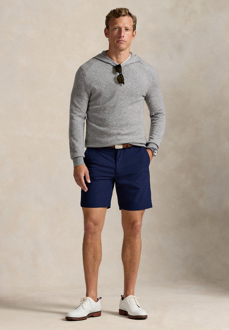 Grauer Kapuzenpullover, marineblaue Shorts und weiße Schuhe. Accessoires umfassen Sonnenbrille und braunen Gürtel. Einfaches Design und lockere Passform.