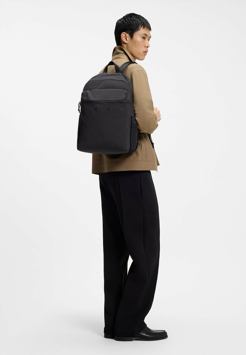 Jeune homme portant une veste beige et un pantalon noir, avec un grand sac à dos noir, debout devant un fond blanc uni.