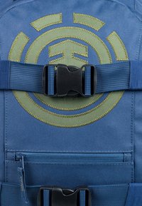 Sac à dos en tissu bleu avec un grand logo circulaire vert brodé, une boucle en plastique noire et une poche zippée à l'avant.