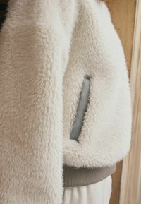 Puszysta, jasno-beżowa krótka kurtka o pluszowej teksturze, z szarym detalem kieszeni i ściągaczem u dołu. Miękki materiał zapewnia ciepło i komfort.