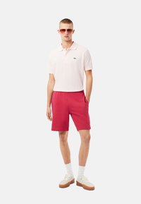 Camiseta polo de color rosa claro con un logo verde, combinada con pantalones cortos rojos brillantes y zapatillas blancas. El modelo lleva gafas de sol y se muestra de manera informal.