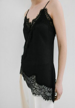 Mujer con camisola negra sin mangas con borde de encaje y dobladillo con borde ondulado, combinada con pantalones blancos, sobre un fondo liso.