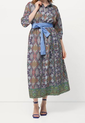 Vrouw die een lange jurk met patroon draagt, een blauwe tailleband en blauwe sandalen met hoge hakken, staand tegen een effen achtergrond.
