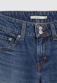 Jean en denim bleu avec fermeture à double bouton métal, rivets aux poches avant, coutures orange et étiquette portant la mention "SUPERLOW 27" sur la ceinture.