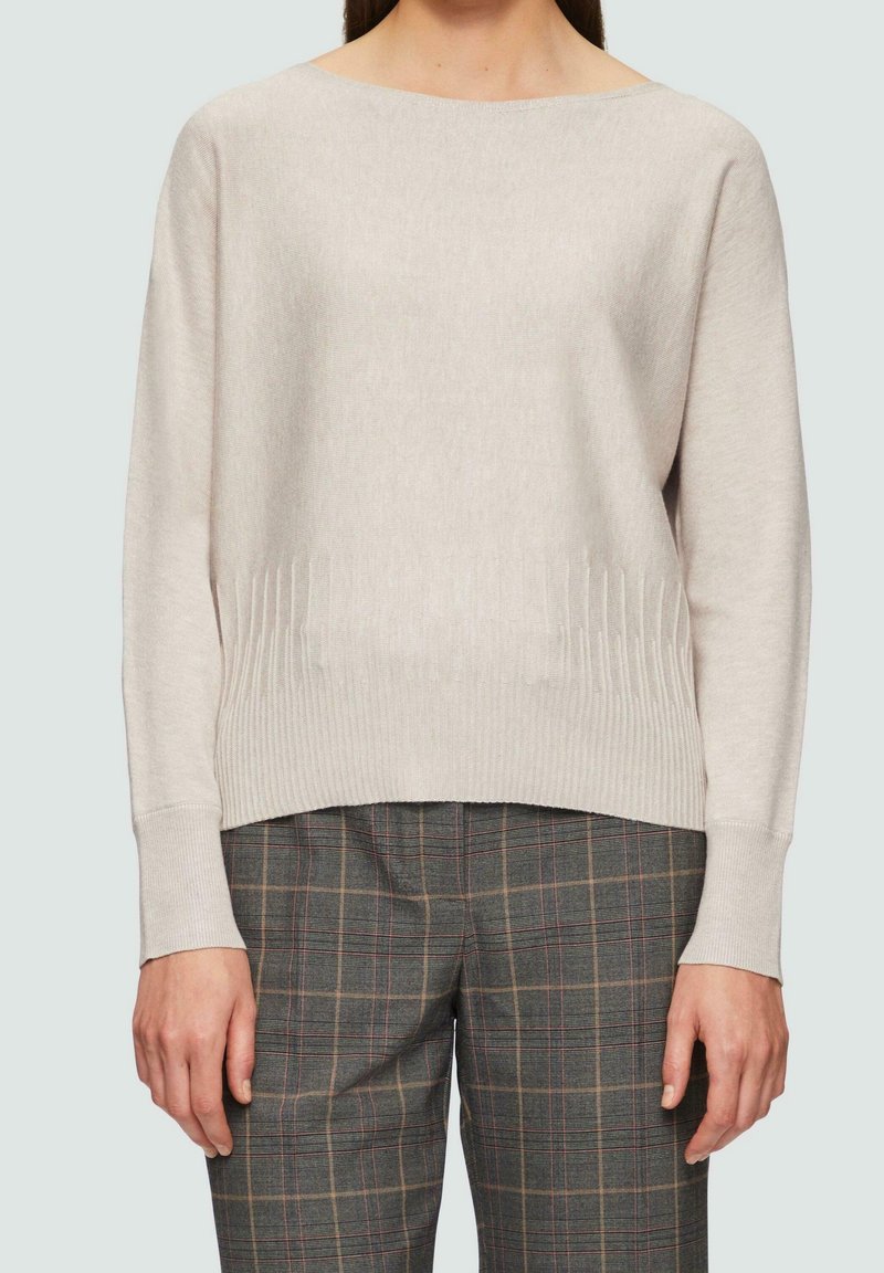 Femme portant un pull léger à manches longues beige clair et un pantalon à carreaux gris, debout devant un fond uni.