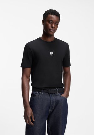 Junger Mann trägt ein schwarzes Kurzarmhemd mit kleinem weißem Logo und dunkelblauen Jeans, steht mit einer Hand in der Hosentasche.