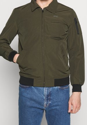 Uomo che indossa una giacca bomber verde oliva con cerniera e polsini neri, jeans blu, una mano nella tasca della giacca, in piedi davanti a uno sfondo neutro.