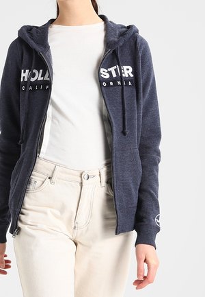 Osoba ubrana w ciemnoniebieską bluzę z kapturem zapinaną na zamek, na której znajduje się biały napis "Hollister California", pod bluzą biała koszulka i jasnobeżowe spodnie.