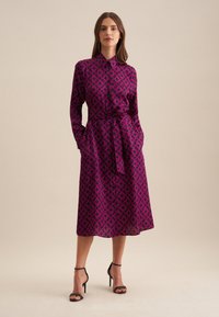 Abito camicia di colore rosa scuro con un motivo geometrico. Presenta un colletto alto, maniche lunghe, una vita definita da una cintura e una lunghezza midi.