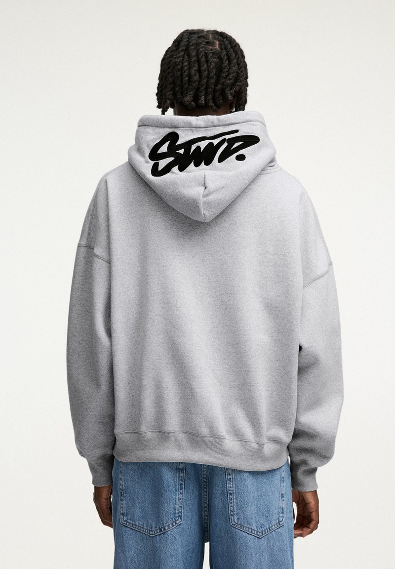 Harmaa huppari, jossa musta logo takana, rento istuvuus, ylisuurikokoinen siluetti sekä resorit hihansuissa ja helmassa.