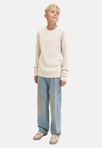 Blonder Junge trägt einen cremefarbenen Kabelstrickpullover, lockere hellblaue Jeans und beige Turnschuhe, steht und sieht leicht nach oben zur Seite.