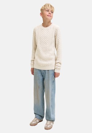 Blonder Junge trägt einen cremefarbenen Kabelstrickpullover, lockere hellblaue Jeans und beige Turnschuhe, steht und sieht leicht nach oben zur Seite.