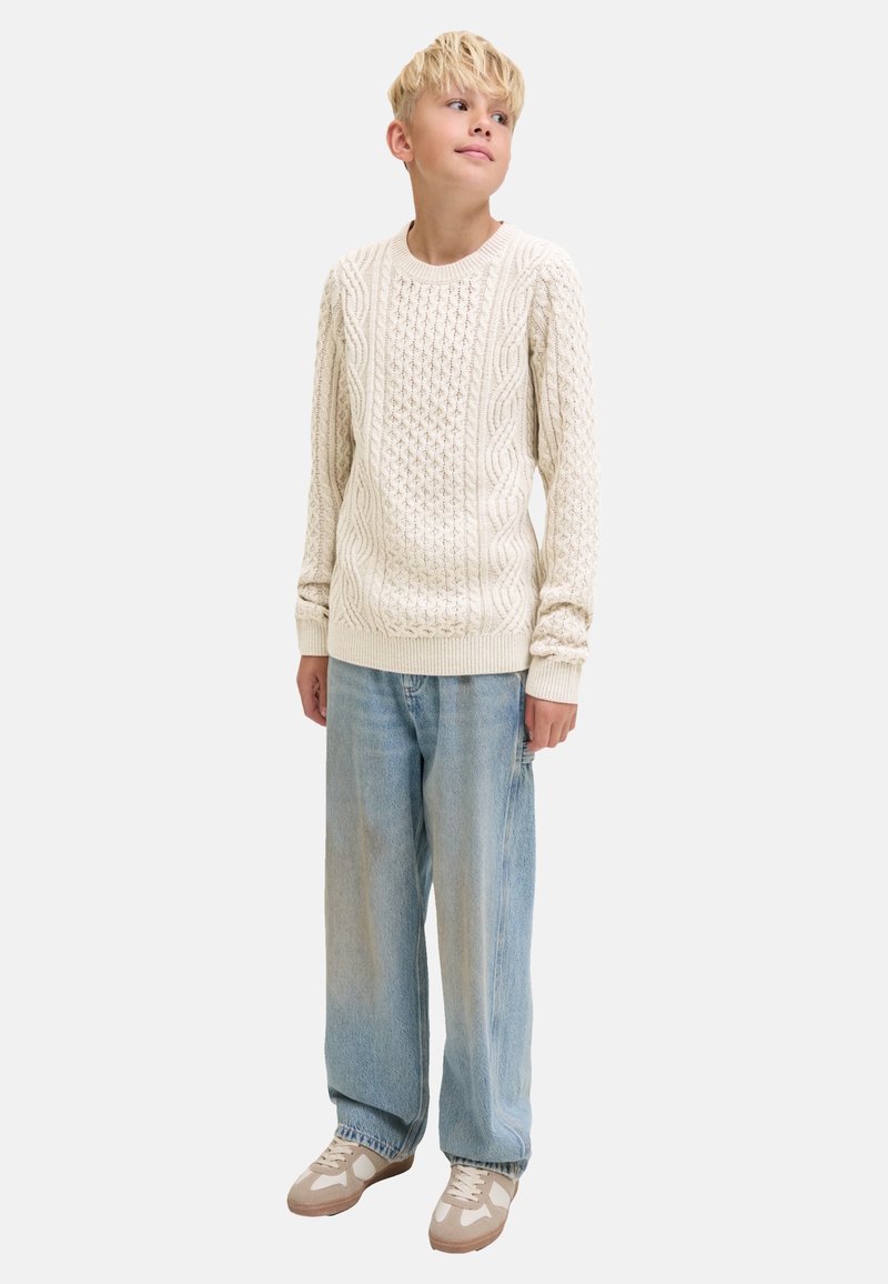 Blonder Junge trägt einen cremefarbenen Kabelstrickpullover, lockere hellblaue Jeans und beige Turnschuhe, steht und sieht leicht nach oben zur Seite.