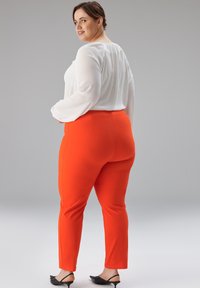 Rote, dehnbare Hose mit figurbetontem Design, kombiniert mit einer weißen Bluse mit langen Ärmeln. Schwarze, spitz zulaufende Schuhe vervollständigen das Outfit.