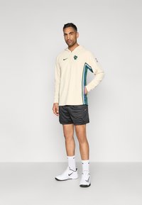 Béžová mikina Nike so zelenými a modrými bočnými pruhmi, čierne sieťované šortky, biele ponožky a biele športové topánky, postavené na jednoduchom pozadí.