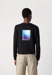 Langärmliger schwarzer Pullover mit einem quadratischen blauen und rosafarbenen Grafik auf der Rückseite, die "calvin klein" zeigt. Aus Baumwollstoff, lockerer Schnitt.