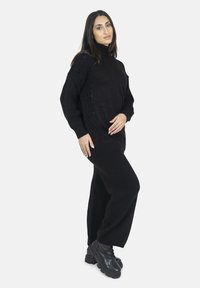 Maglione nero a maglia con collo alto e polsini a coste, abbinato a pantaloni neri a gamba larga e robusti stivali neri.
