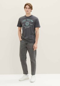 Dunkelgraues Baumwoll-T-Shirt mit bedrucktem Text in Blau und Weiß, kombiniert mit dunklen Jeans und weißen Sneakers. Einfaches Rundhalsdesign.