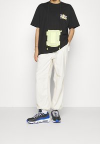 Zwart T-shirt met korte mouwen en logo, witte wijde broek, en zwarte sneakers met blauwe accenten. Een lichtgroene crossbody tas hangt aan een strap.