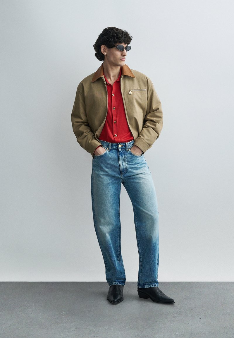 Man draagt een beige jas over een rood shirt, lichtblauwe spijkerbroek, zwarte laarzen en zwarte zonnebril, staand met handen in de zakken tegen een effen achtergrond.