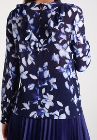 Navybluse med hvidt og blåt blomsterprint, med en flæsekantet krave og knaplukning bagpå, med lange ærmer.