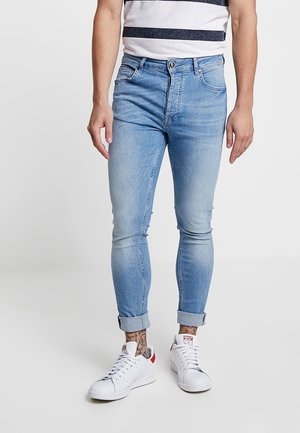 Man wearing light blue skinny jeans rolled at the ankles, white sneakers with red detail, and a white and navy striped shirt.