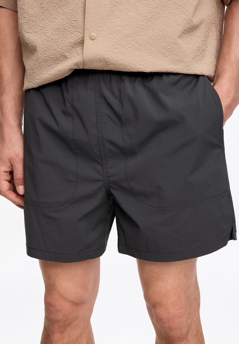 Homme portant un short noir à taille élastique et une chemise beige texturée à manches courtes, les mains reposant près des poches, debout sur un fond blanc.