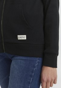 Felpa nera con zip realizzata in tessuto a costine, caratterizzata da due tasche laterali e una piccola etichetta bianca nella parte anteriore in basso. Abbinata a jeans blu.