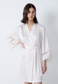 Robe in seta di un delicato crema con vita cinturata, maniche a kimono e pizzo decorativo sui polsini. Texture liscia e design minimalista.