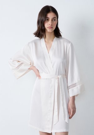 Peignoir en satin de couleur crème clair avec une taille ceinturée, des manches kimono et des accents en dentelle sur les poignets. Texture lisse et design minimaliste.
