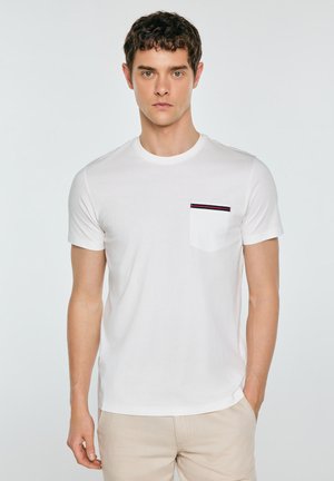 Młody mężczyzna w białej koszulce z krótkim rękawem z kieszenią ozdobioną paskami, stojący z jedną ręką w kieszeni beżowych spodni.