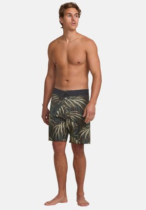 Manlig modell som bär mörka badshorts med ett grönt och beige palmblads mönster, snörning i midjan och mjukt tyg. Inga skor bärs.