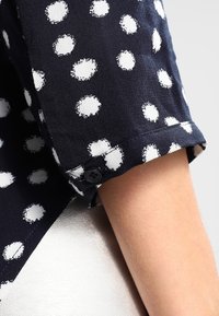 Marineblaue Bluse mit weißen Punkten, featuring umgekrempelte Ärmel mit einem schwarzen Knopfdetail und einer glatten Stoffstruktur.