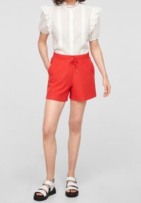 Rode geribde shorts met een elastische tailleband en een trekkoord. Gecombineerd met een witte blouse met rimpelende mouwen en kantafwerking. Zwarte sandalen.