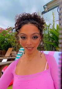 Long-sleeve, fitted top in sheer light pink fabric with small embellishments, paired with layered gold necklaces, smiling outdoors.