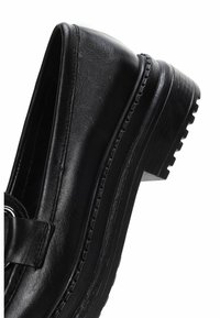 Zapato de cuero negro con punta redondeada y suela gruesa y texturizada, que presenta una correa decorativa y un detalle metálico a lo largo del borde superior.