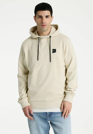 Jeune homme portant un sweat à capuche beige avec des cordons noirs et un petit logo noir, associé à un jean bleu clair, debout devant un fond blanc.
