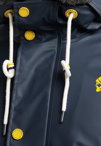 Marinblå vattentät jacka med vita dragsnören, gula detaljer och logotyp. Har en slät textur och runt gula logotypknappar på huvan.