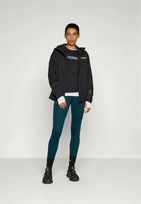 Személy áll fekete Terrex dzsekiben és pólóban, türkiz leggingsben, fekete zokniban és fekete sportcipőben egy egységes háttér előtt.