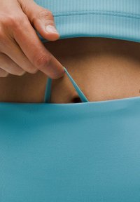 Ruka tahá světle modrou látku, čímž odhaluje tenký popruh pod ní. Důraz je kladen na textury a akci úpravy oblečení.