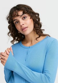 Cardigan bleu en tissu tricoté doux, avec un col rond, une fermeture à boutons à l'avant et des manches longues avec des poignets côtelés.