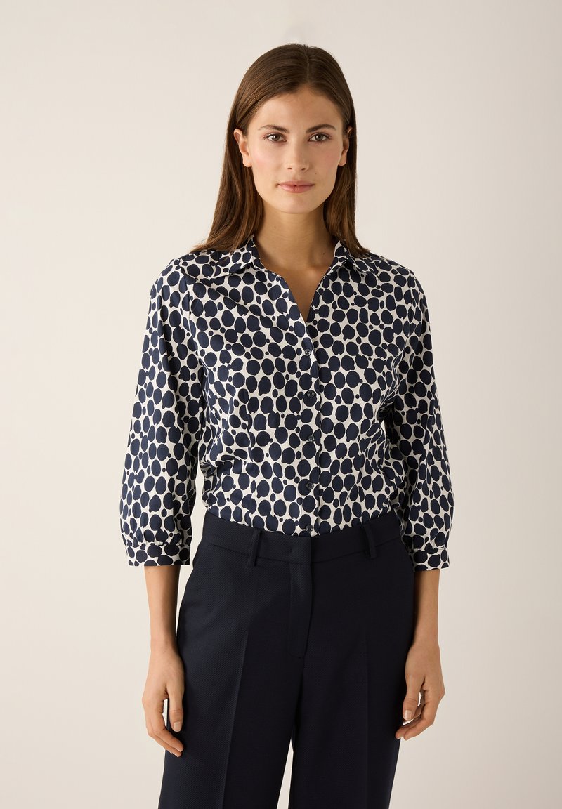 Navy and white polka dot button-up shirt with a collar, three-quarter sleeves, and a relaxed fit, paired with dark tailored pants.