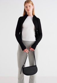 Schwarze Zip-Jacke über einem weißen Oberteil, kombiniert mit grauen Hosen. Halten einer kleinen schwarzen Umhängetasche in gebogener Form mit langem Tragegurt.