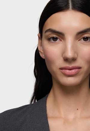 Donna con capelli lunghi e scuri che indossa piccoli orecchini a bottone quadrati e un top grigio scuro, espressione neutra, su uno sfondo chiaro e uniforme.