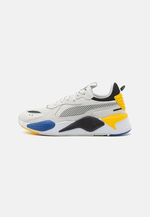 Zapatilla gris clara que combina materiales de malla y ante, con acentos negros y detalles coloridos en azul y amarillo en el talón y la suela.