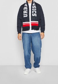 Granatowa pikowana kurtka bomberka z ribbanym mankietami, biały t-shirt, jasnoniebieskie dżinsy w szerokim kroju i czarny szal w czerwono-białe paski. Białe sneakersy.