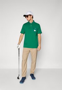 Polo verde cu un buzunar pe piept, pantaloni bej, șapcă albă, ochelari de soare și mănușă de golf, ținând un club de golf, pe un fundal simplu.