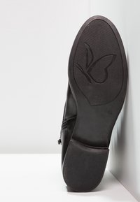 Chaussure en cuir noir avec une semelle en caoutchouc texturée, présentant un logo de papillon embossé sur le dessous et un design lisse et courbé.