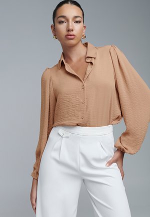Beige kaulushirt, jossa on puffit pitkät hihat, yhdistetty valkoisiin korkeaselkäisiin housuihin, joissa on laskokset ja nappiyksityiskohta.